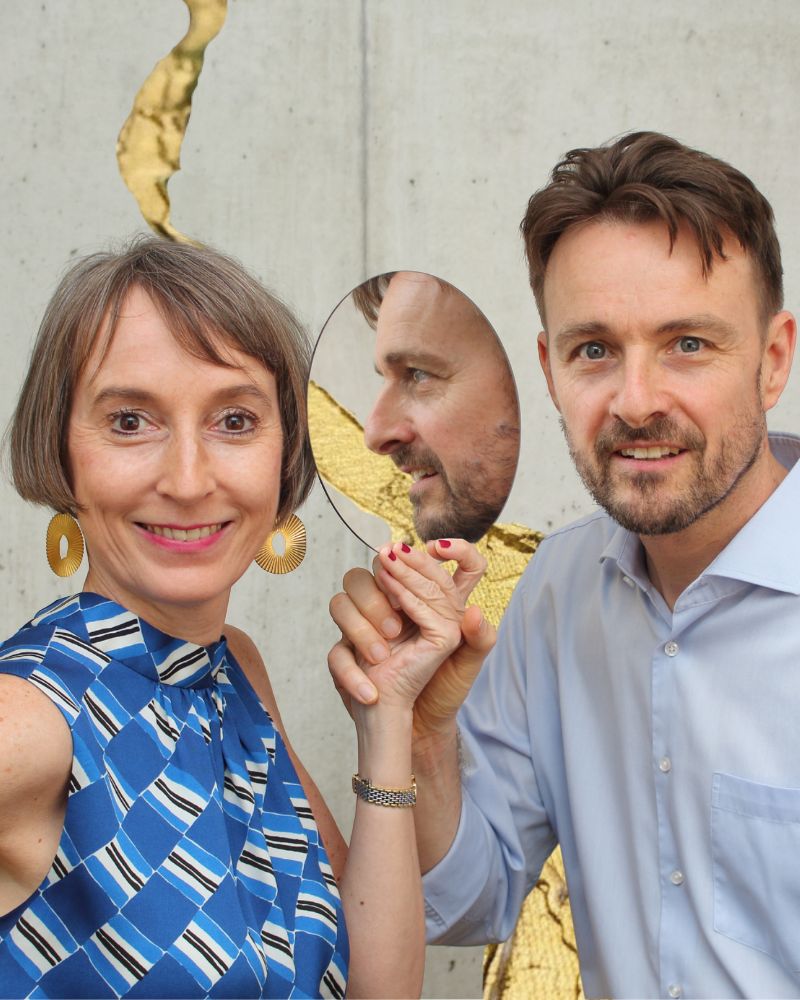 Frau und Mann halten zusammen einen Spiegel in den Händen und blicken den Betrachtenden an